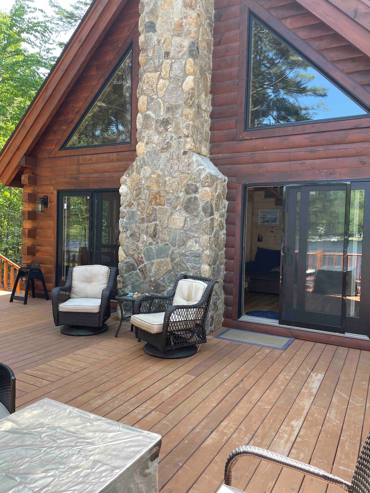 A spacious wooden deck is featured in front of a log cabin, with a stone chimney rising between large glass windows. Two comfortable chairs, one cushioned and one woven, are arranged to create a relaxing seating area, complemented by the natural surroundings.