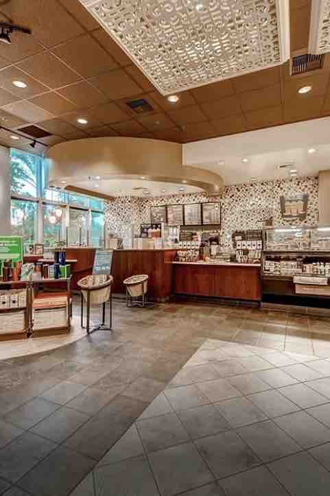 A modern café space presents a curved counter adorned with an array of baked goods and beverages. Wall decor features intricate patterns, while natural light enters through large windows. Nearby seating is arranged with high chairs and small tables, creating a welcoming atmosphere for guests.