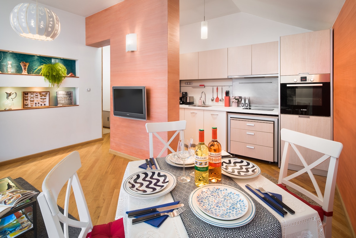A dining area is showcased with a round table set for four, featuring patterned plates and glasses. In the background, a modern kitchen is visible, equipped with sleek appliances and a range of utensils, complemented by warm wooden flooring and colorful accent walls.
