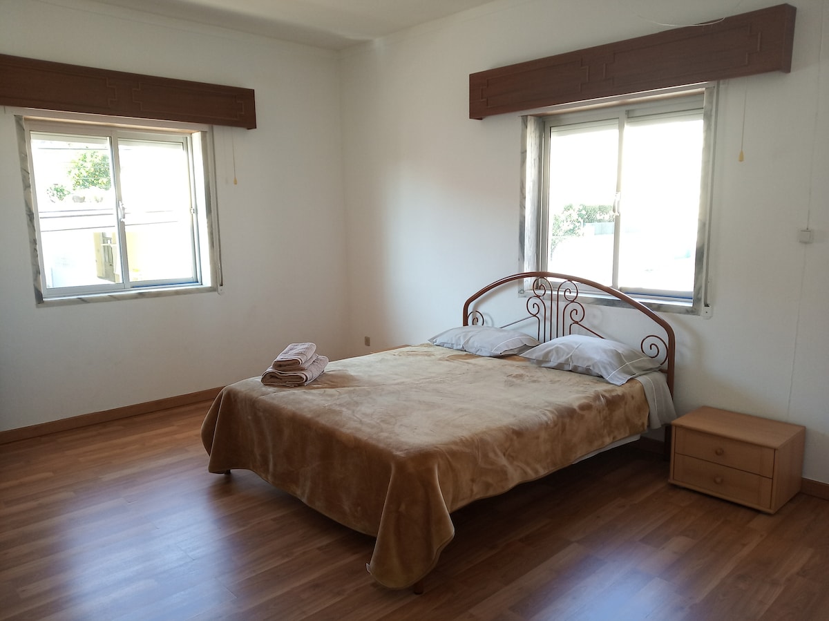 A room featuring a spacious double bed covered with a light brown blanket. Two windows allow natural light to illuminate the space, complemented by a small wooden nightstand beside the bed. The room is equipped with wooden flooring and has a minimalist design.