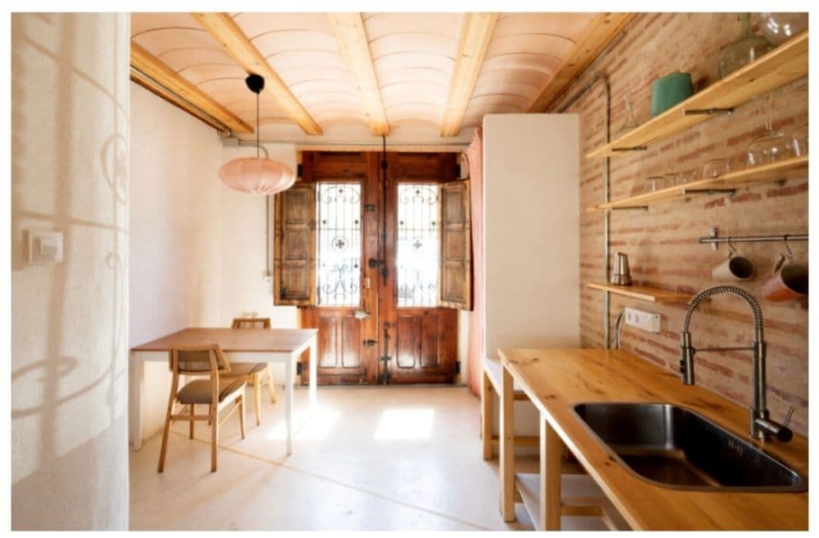 An inviting kitchen area features wooden shelves displaying glassware and a light pink pendant light. A wooden table with two chairs stands opposite a kitchen counter with a stainless steel sink, complemented by rustic brick walls and double wooden doors adorned with intricate ironwork.