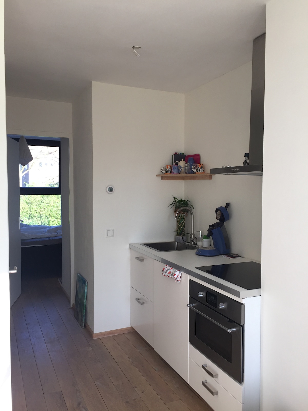 A compact kitchen area is visible, featuring a sleek black countertop and modern appliances, including a stove and oven. Shelves hold kitchenware, while a window allows natural light to enter. Light wood flooring enhances the open feel of the space.