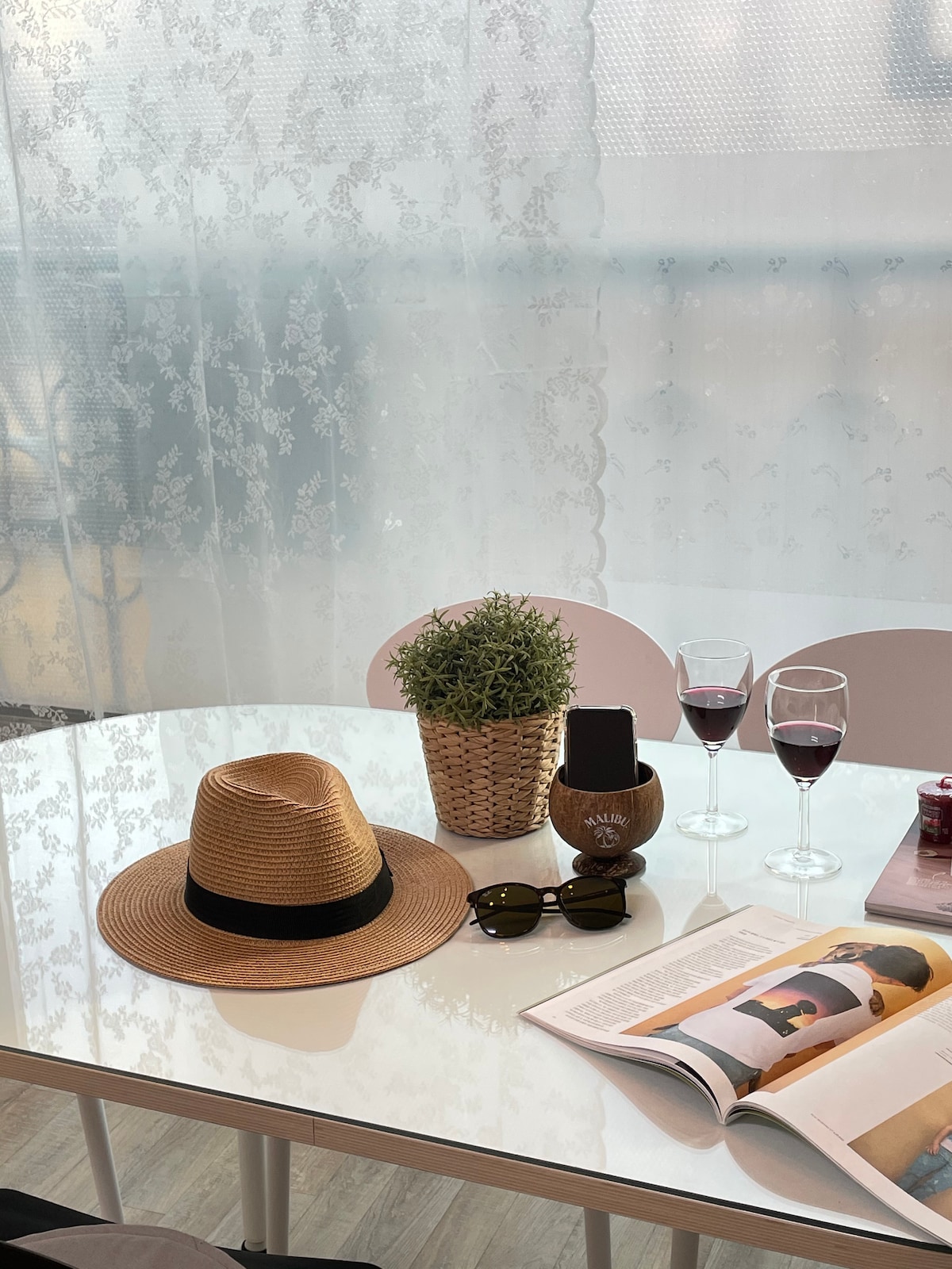 A bright and airy dining table is set with a potted plant and two glasses of red wine. A sun hat and sunglasses are placed beside an open magazine, creating a relaxed atmosphere illuminated by natural light filtering through a lace curtain.