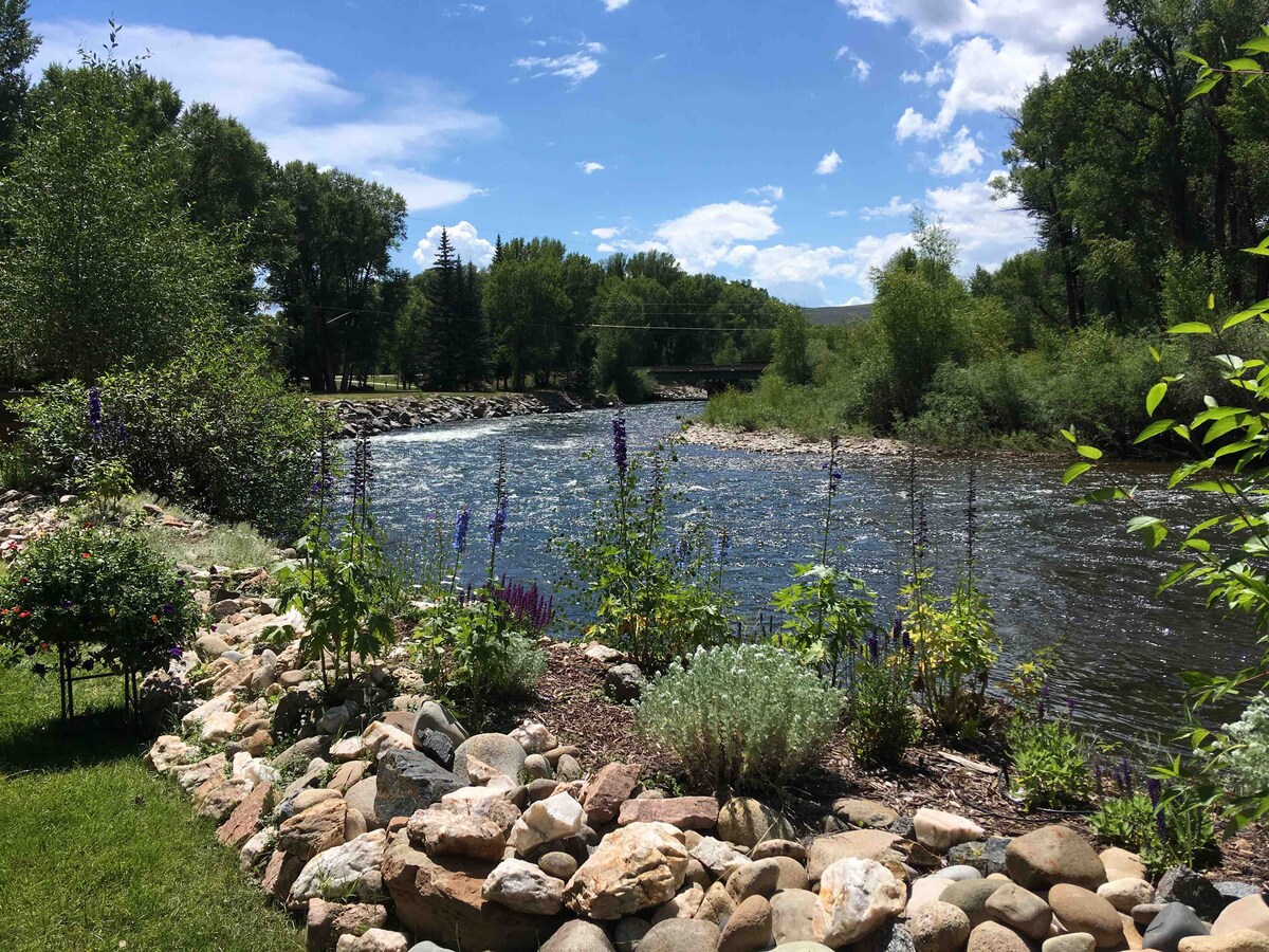 The scenic view showcases the Gunnison River flowing gently alongside a landscaped area filled with colorful flowers and green foliage. Smooth stones line the bank, leading to the shimmering water. Lush trees and blue skies frame the tranquil river, creating a peaceful outdoor setting.