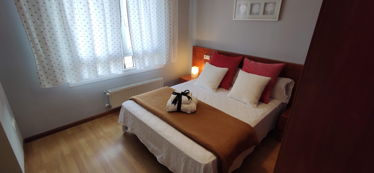 A cozy bedroom is presented with a spacious bed featuring white linens and complementary pillows in shades of red and white. Natural light filters through a window adorned with polka dot curtains, illuminating the warm wooden flooring and a bedside table with a soft lamp.