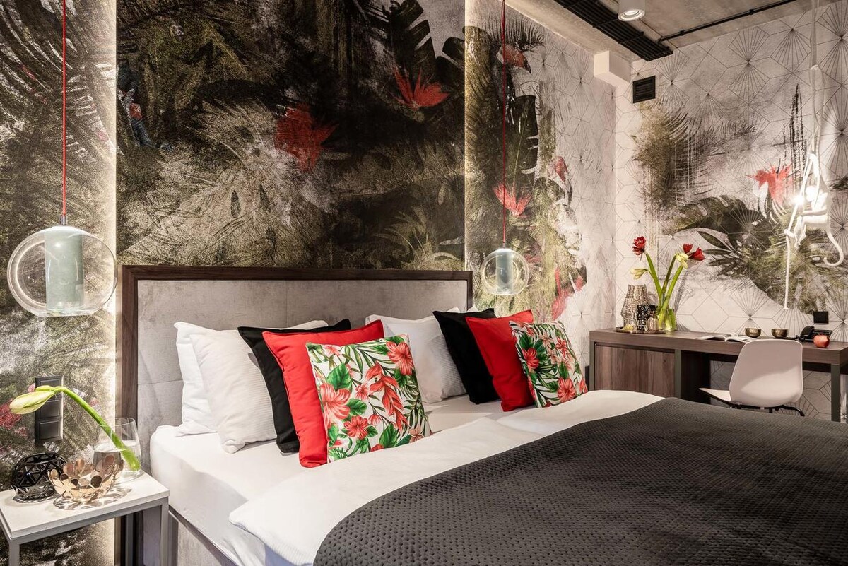 A modern room is filled with tropical-themed wallpaper featuring lush greenery and vibrant flowers. A spacious bed is dressed with decorative pillows and a dark blanket. A sleek desk and chair are positioned nearby, along with soft lighting fixtures contributing to the overall ambiance.