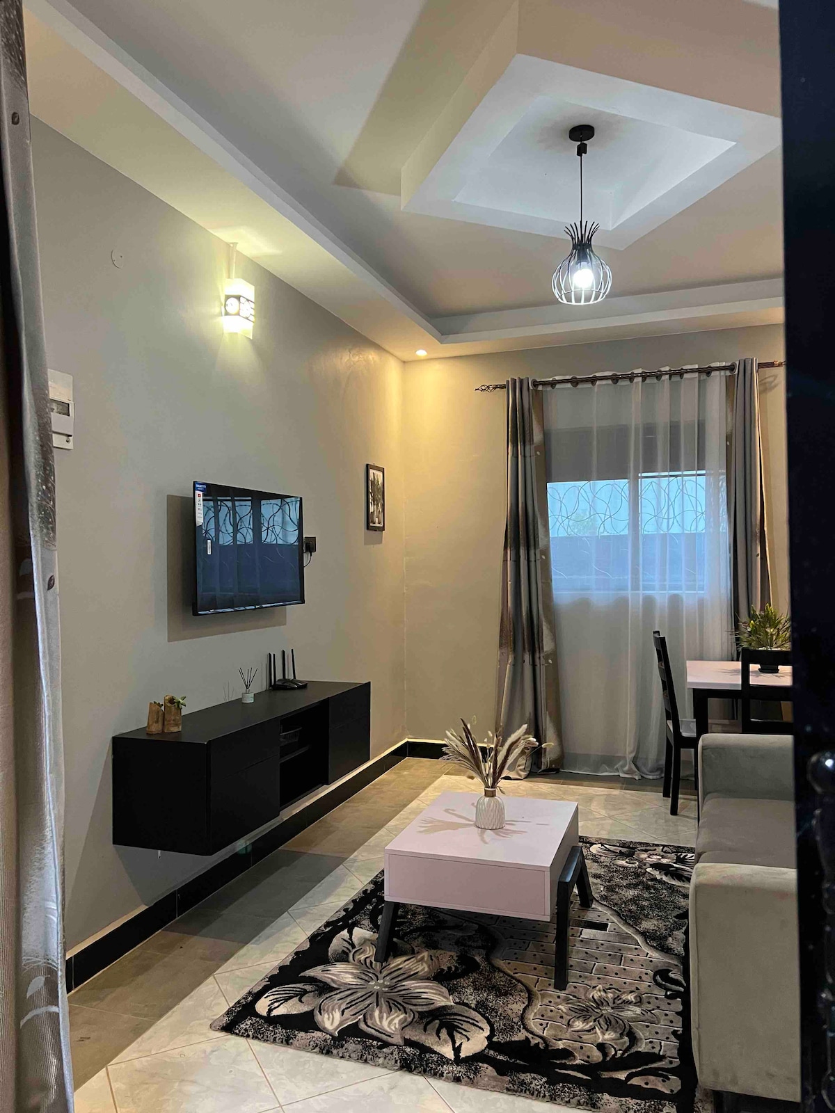 A living area features a sleek black television mounted on a light gray wall. A small coffee table with a floral design sits on a patterned rug, surrounded by a comfortable sofa. Natural light filters through sheer curtains at a window.