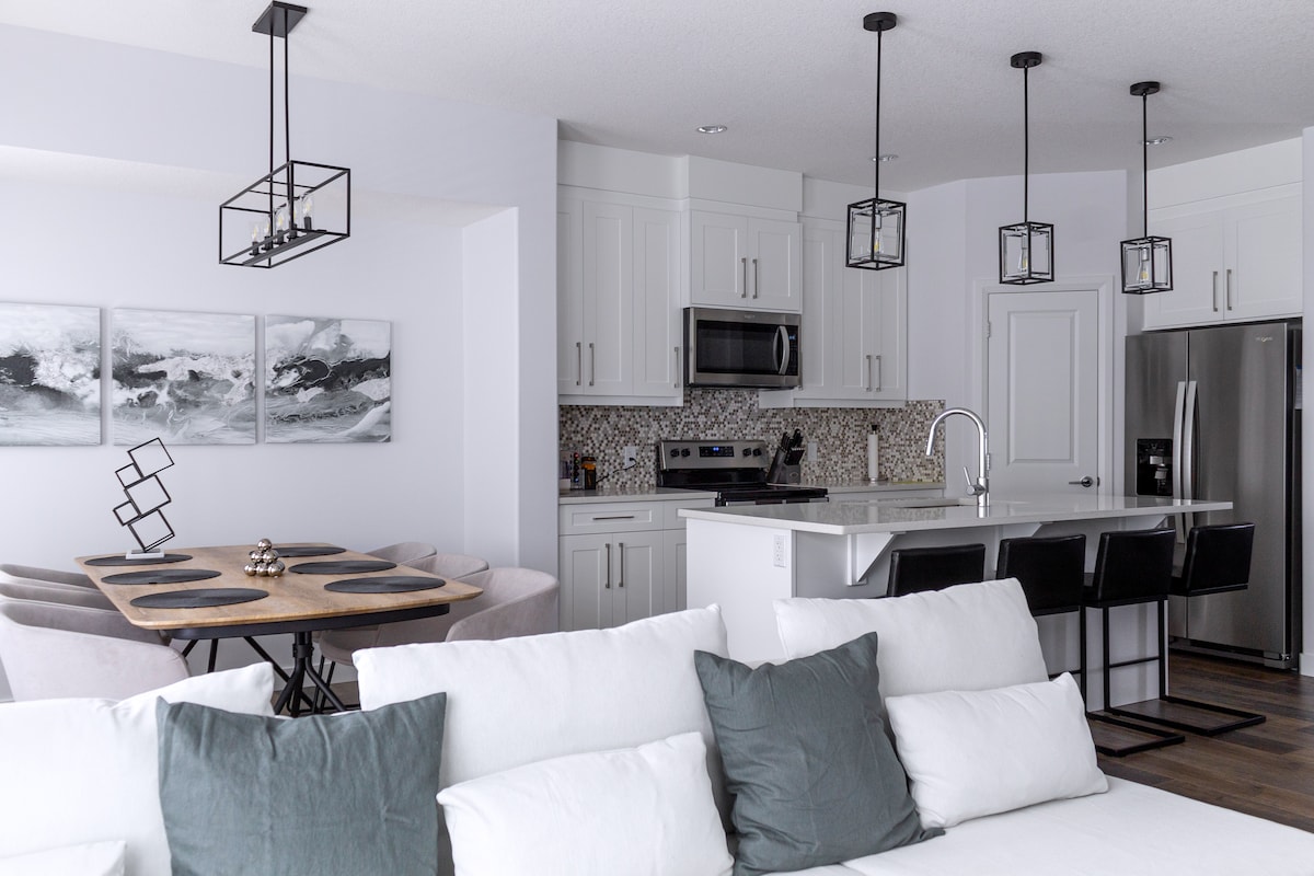 An open-concept kitchen and dining area is showcased. The kitchen features modern appliances, including a microwave and stainless steel refrigerator. A long island bar with stools is visible, alongside a dining table set for four. Unique light fixtures hang from the ceiling, complementing minimalistic decor.