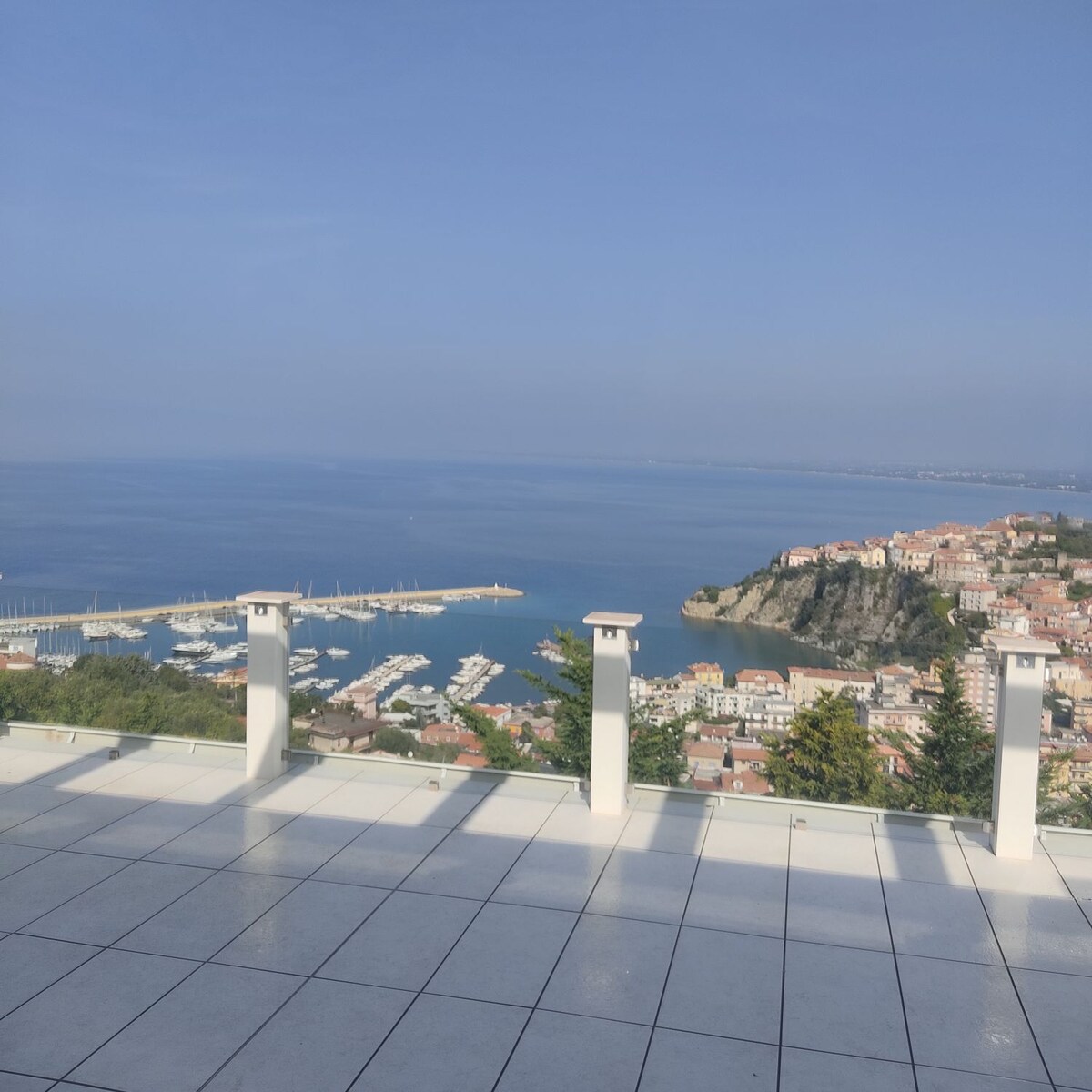 A large terrace offers a panoramic view of the coastline and marina, with calm blue waters extending to the horizon. White pillars frame the terrace, and sunlight casts gentle shadows across the tiled floor, enhancing the serene setting.