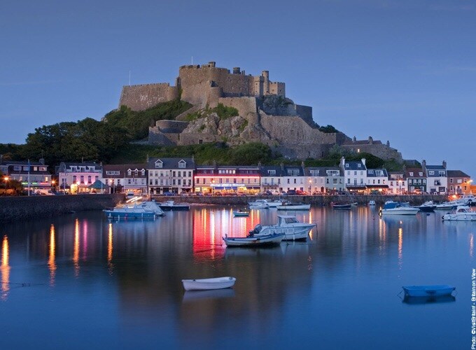 A coastal scene is captured at dusk, showcasing a historic castle atop a hill illuminated by twilight. The calm waters reflect various boats and the colorful lights of nearby buildings, creating a serene atmosphere along the harbor.