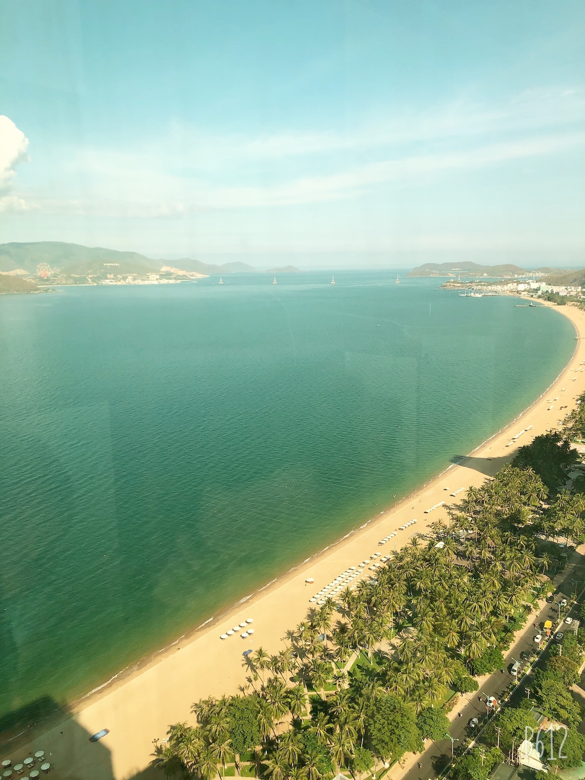 A panoramic view of the coastline captures the serene blue waters meeting the sandy beach. Lush palm trees line the shore, while distant hills and the city skyline create a scenic backdrop under a clear sky.