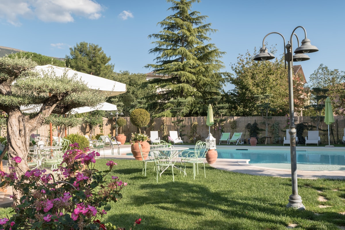 A serene garden setting is depicted, featuring a clear pool surrounded by neatly arranged lounge chairs. Ornamental plants and vibrant flowers add color to the space. A stone path leads to the pool area, and shade is provided by large umbrellas and mature trees.