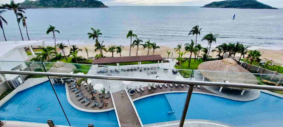 A panoramic view of the coastline from a balcony showcases a beautiful sandy beach bordered by palm trees. Two swimming pools can be seen on the property, surrounded by lounge chairs and shaded areas for relaxation.