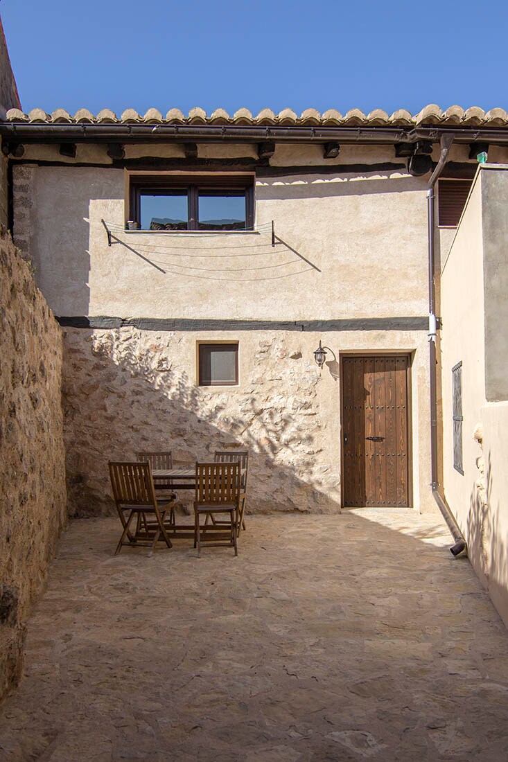 A private outdoor patio is depicted, featuring a textured stone surface. Wooden tables and chairs are arranged, providing a space for relaxation. The building's rustic façade contrasts with the bright sky, and a wooden door is visible, contributing to the inviting atmosphere.