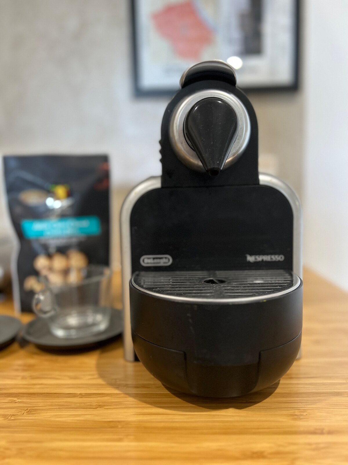 A sleek black coffee machine sits prominently on a wooden countertop. A glass cup rests nearby, accompanied by a bag of coffee pods, suggesting a convenient coffee-making setup. The background includes a map, adding a touch of local character.