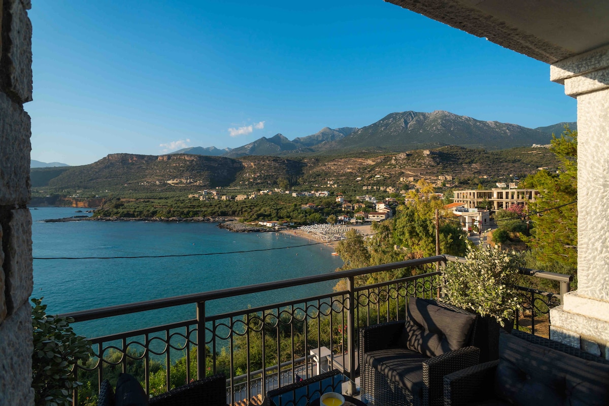 A panoramic view of the sea and coast is captured from a balcony. Lush green hills rise in the background, while a quaint beach is visible below, dotted with umbrellas. A comfortable seating arrangement is positioned on the balcony, inviting relaxation.