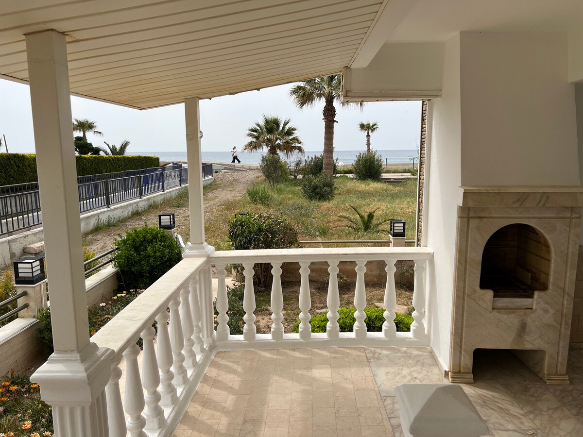 A covered terrace with marble flooring provides a view of the grassy area leading to the beach. Palm trees and plants are visible in the surroundings, with a pathway leading toward the sand. A stone fireplace is unobtrusively positioned along the terrace.