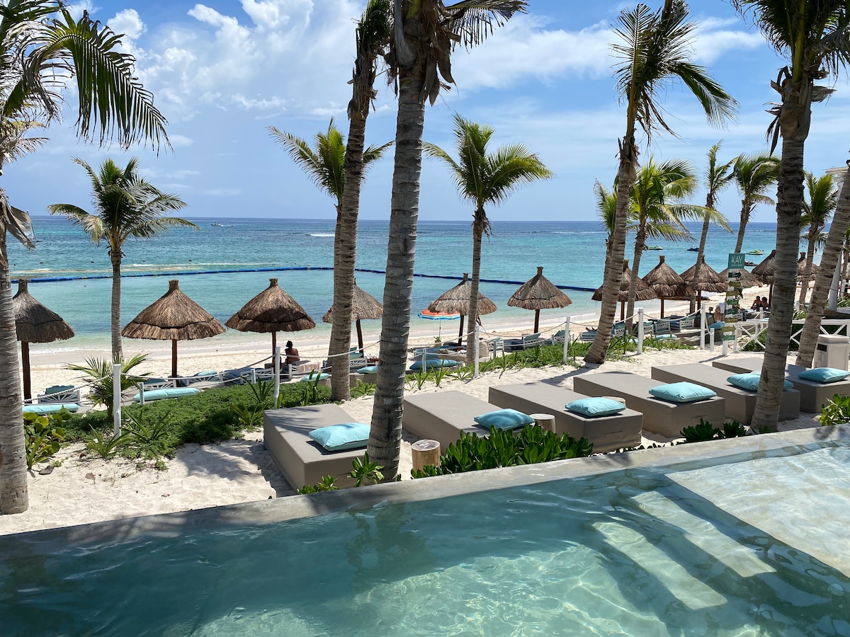 A view of the beach features sandy shores lined with sunbeds and thatched umbrellas. Palm trees sway gently, framing the inviting turquoise waters of the sea. An infinity pool with a clear reflection mirrors the serene landscape, offering a calming atmosphere.