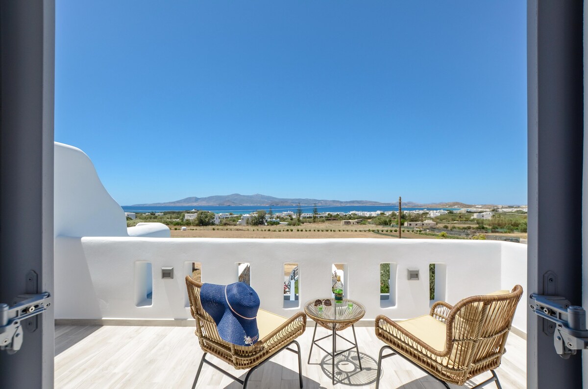 A spacious veranda with a light wooden floor features two comfortable chairs and a small round table. A blue hat rests on the table, providing a relaxed ambiance. The expansive view showcases the sea and distant landscapes under a clear blue sky.