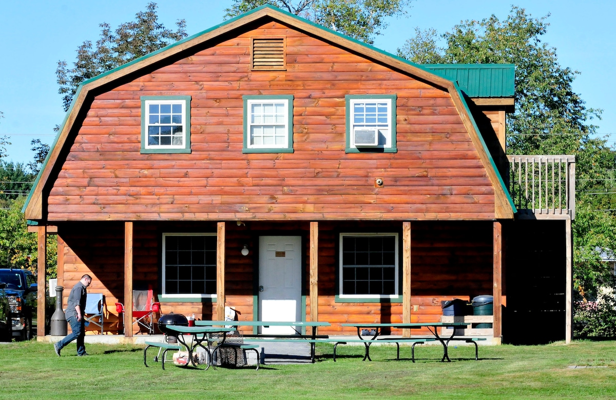 The two-story cabin features a rustic wooden facade, complemented by a green roof. A central door is flanked by large windows. An outdoor seating area with picnic tables is visible on the lawn, surrounded by a well-maintained grassy area.