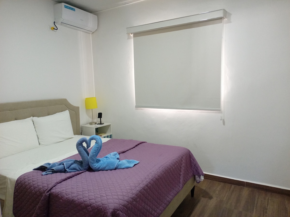 A well-lit bedroom features a spacious bed dressed in soft linens. A bedside table with a lamp complements the decor. A cozy blanket is draped across the foot of the bed, with a pair of towel swans arranged elegantly on top.