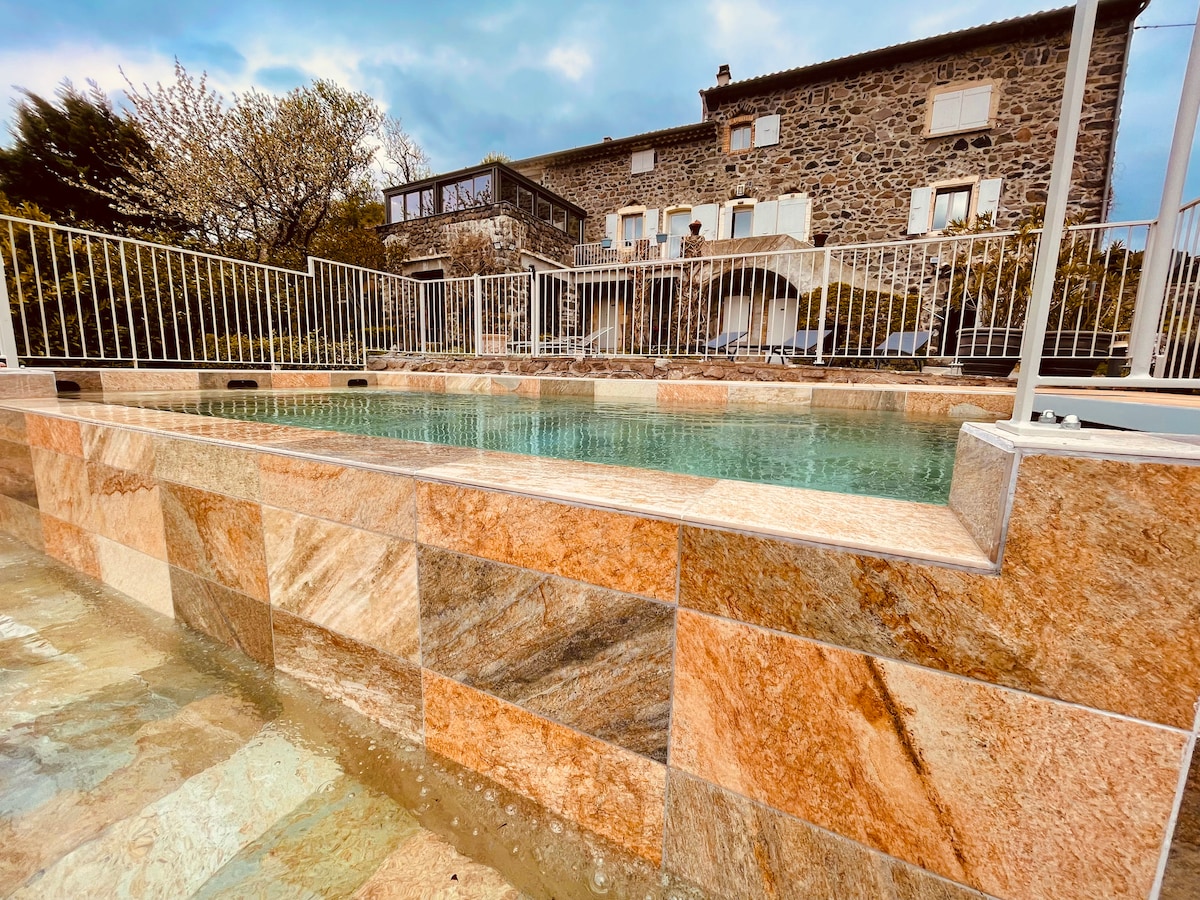 A tranquil infinity pool is surrounded by a stone terrace, featuring a mixture of warm colors on the pool's edge. The rustic stone house in the background provides a contrasting texture against the landscape. Lush greenery can be seen in the surroundings.