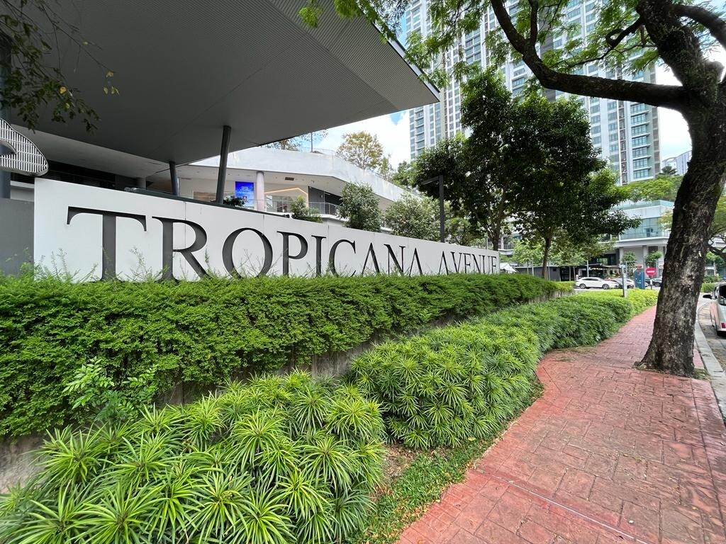 The entrance to Tropicana Avenue is marked by a large sign, surrounded by low shrubs and neatly maintained greenery. A paved walkway leads towards the building, flanked by trees providing shade. The modern architecture of nearby structures can be seen in the background.