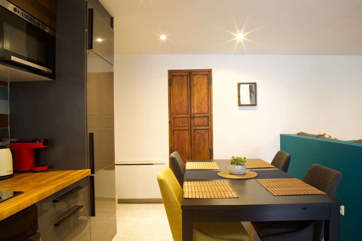 A modern kitchen and dining area features a dark wooden table set for six, accompanied by comfortable, upholstered chairs in varying colors. A small plant sits at the center, with a contemporary kitchen space visible in the background, including cabinetry and appliances.