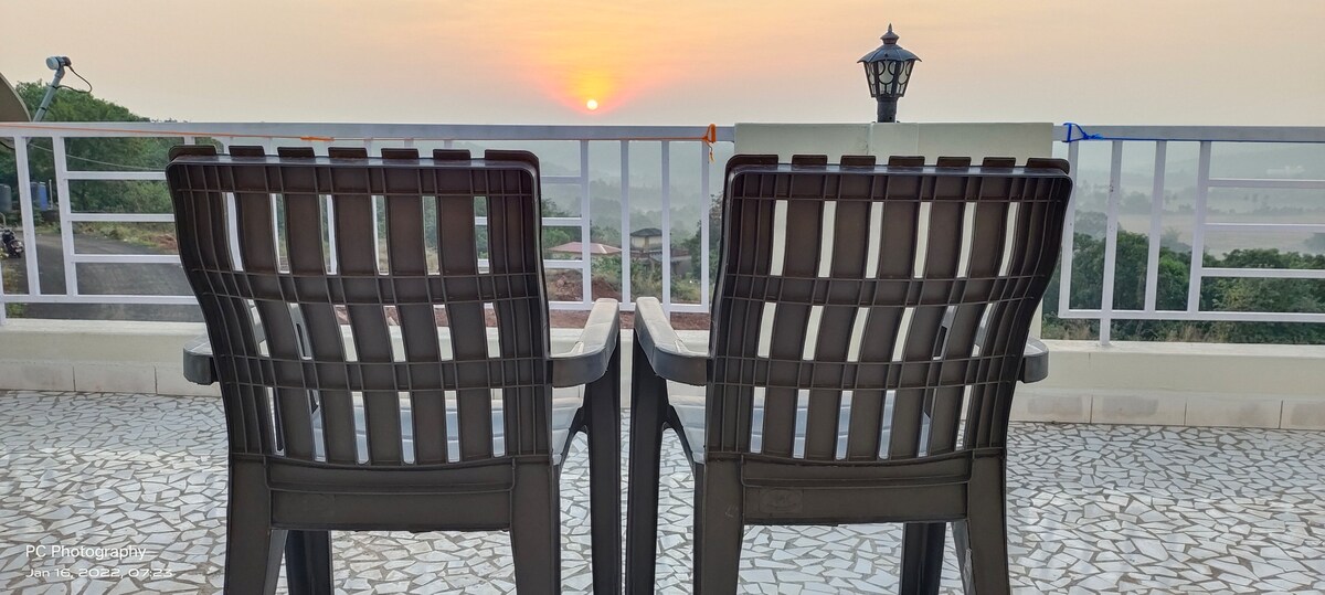 Two black plastic chairs are positioned on a balcony, facing a scenic view of the sunrise over distant hills. The balcony features a white railing and is paved with small, light-colored stones, creating a serene outdoor space.
