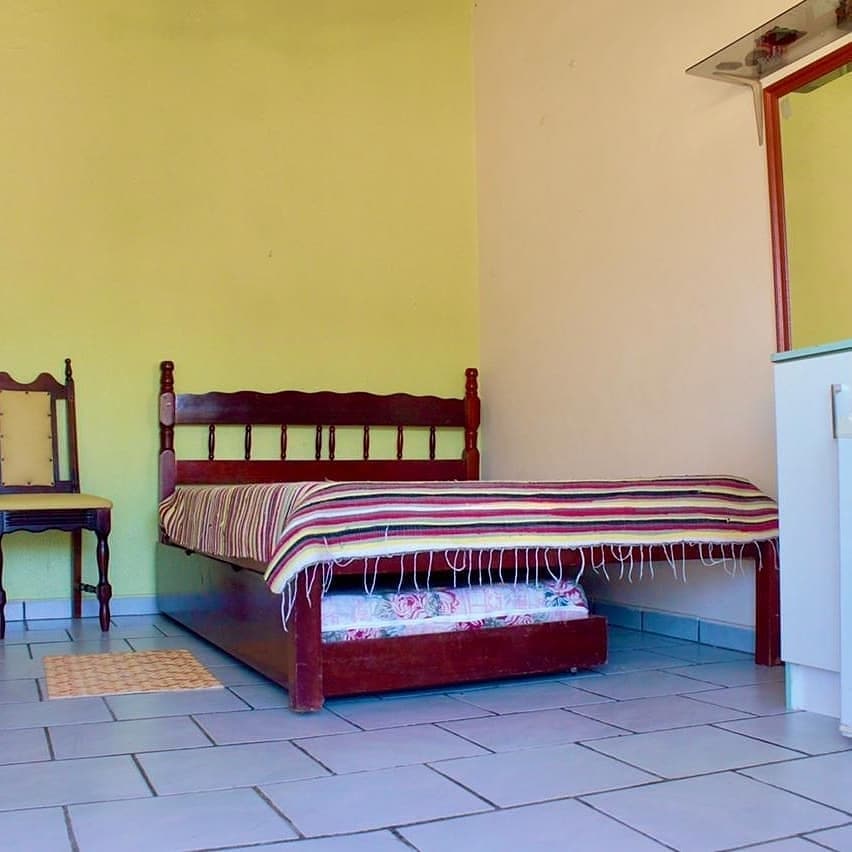 A simple sleeping area features a wooden bed with a striped blanket. A small chair is positioned nearby. Light-colored walls provide a bright backdrop, while tiled flooring contributes to the room's clean appearance.