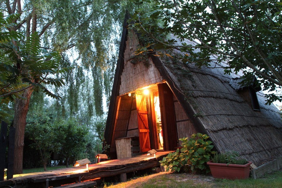 A charming structure features a thatched roof with an A-frame design. Warm light illuminates the entrance, inviting guests inside. Surrounded by trees and green foliage, the pathway leads to the front door, enhancing the serenity of the natural setting.