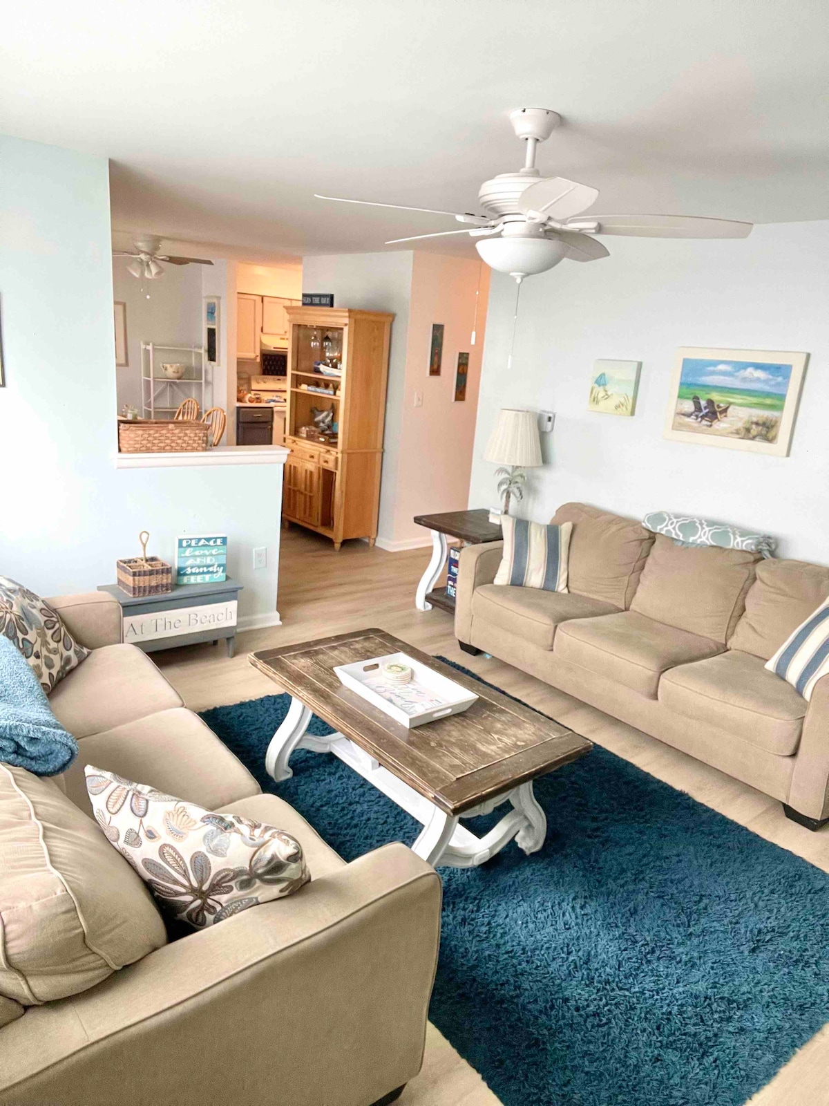 A comfortable living area features two sofas and a coffee table set on a plush blue rug. Light from a ceiling fan creates a welcoming ambiance. The walls are adorned with decorative art, and a view of the adjoining kitchen is visible in the background.