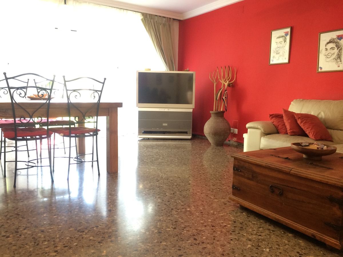An inviting living space features a combination of a wooden dining table with metal chairs and a comfortable sofa. A large television is positioned against a bright red wall, complemented by decorative items and a wooden coffee table on polished flooring.