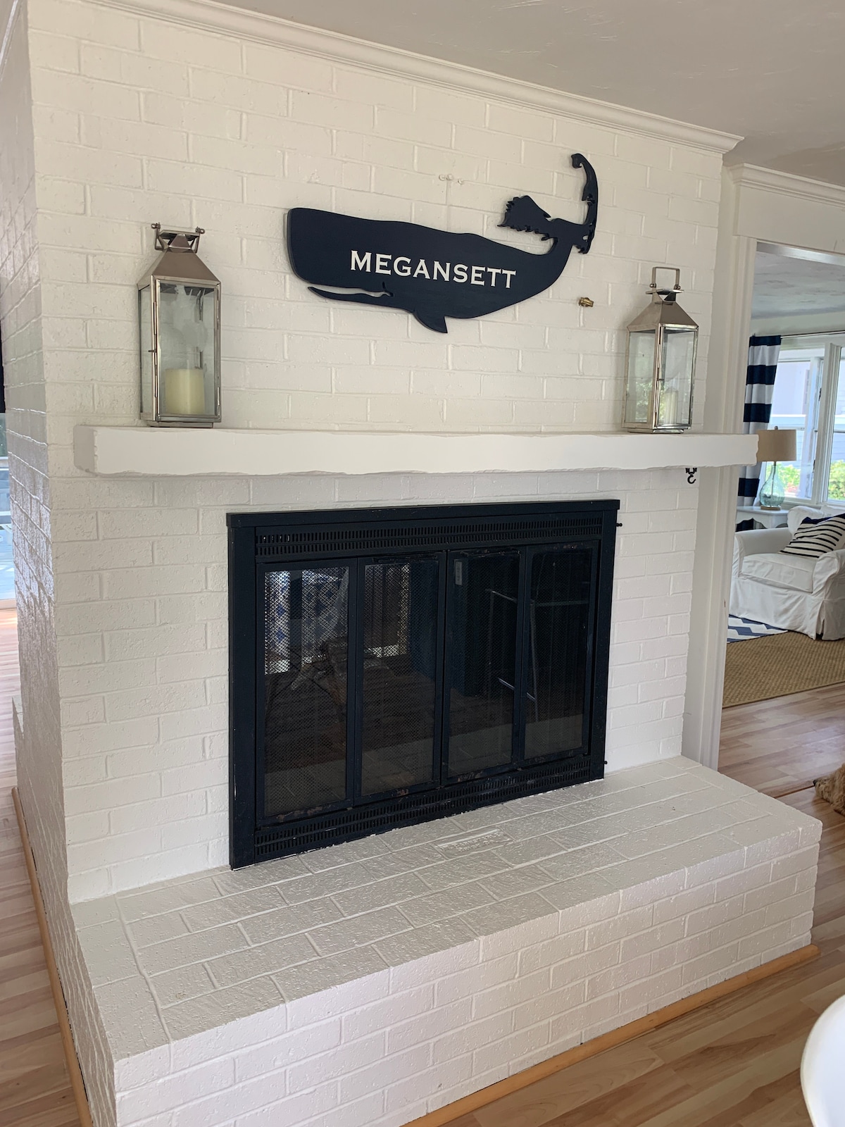 A white brick fireplace is centered in the image, featuring a black grate and a concrete mantel. Two glass lanterns are mounted on either side, enhancing the space. Above the fireplace, a wooden sign displays 'MEGANSSETT,' complementing the neutral tones of the room.
