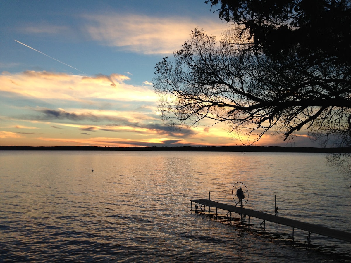 A tranquil scene of Torch Lake at sunset is depicted, highlighting the calm water's surface reflecting vibrant colors. A dock extends into the lake, accompanied by a tree silhouette framing the view, evoking a serene atmosphere. Wispy clouds add texture to the sky.