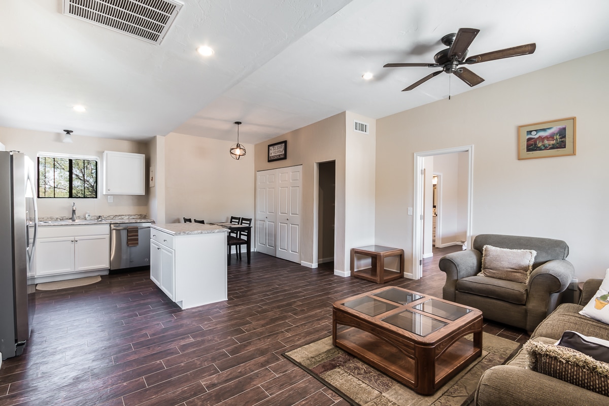 The open living area is designed with natural light highlighting the wood-style flooring. A spacious seating arrangement includes a light grey sofa and a wooden coffee table. The kitchen features modern appliances, while a dining area with chairs is visible in the background.