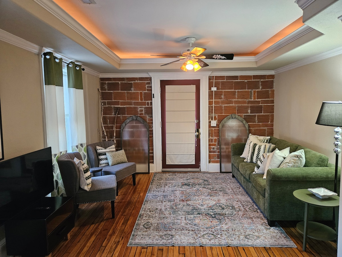 A living area features a green sofa, accompanied by two accent chairs beside a flat-screen television. Natural light enters through large windows adorned with green and white curtains, while brick accents and wooden flooring enhance the room's character.