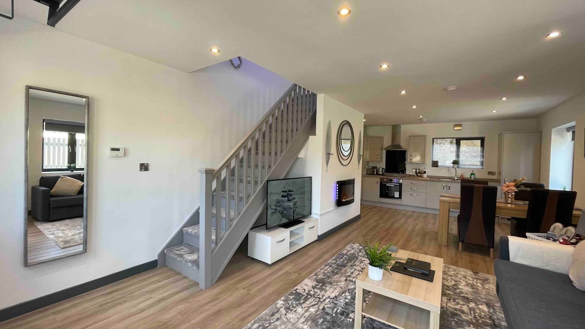 An open-plan living area features a comfortable sofa, a staircase leading to the upper floor, and a Smart TV mounted on the wall. Natural light fills the space through large windows, while a dining table and kitchen fixtures are visible in the background.