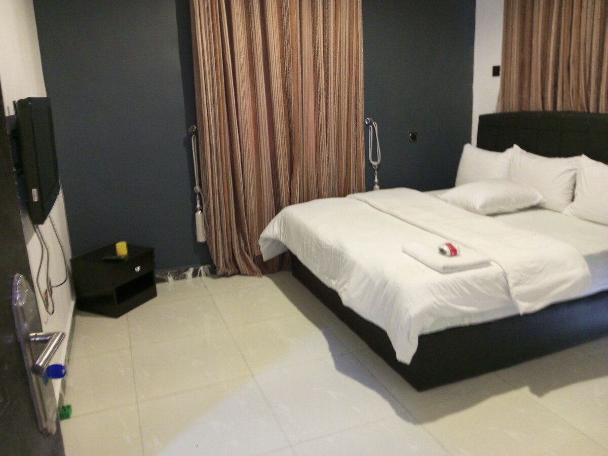 A well-appointed room features a spacious bed with crisp white linens and a variety of pillows. Soft neutral tones are complemented by elegant curtains. A flat-screen television is mounted on the wall opposite the bed, and a small bedside table is located nearby.