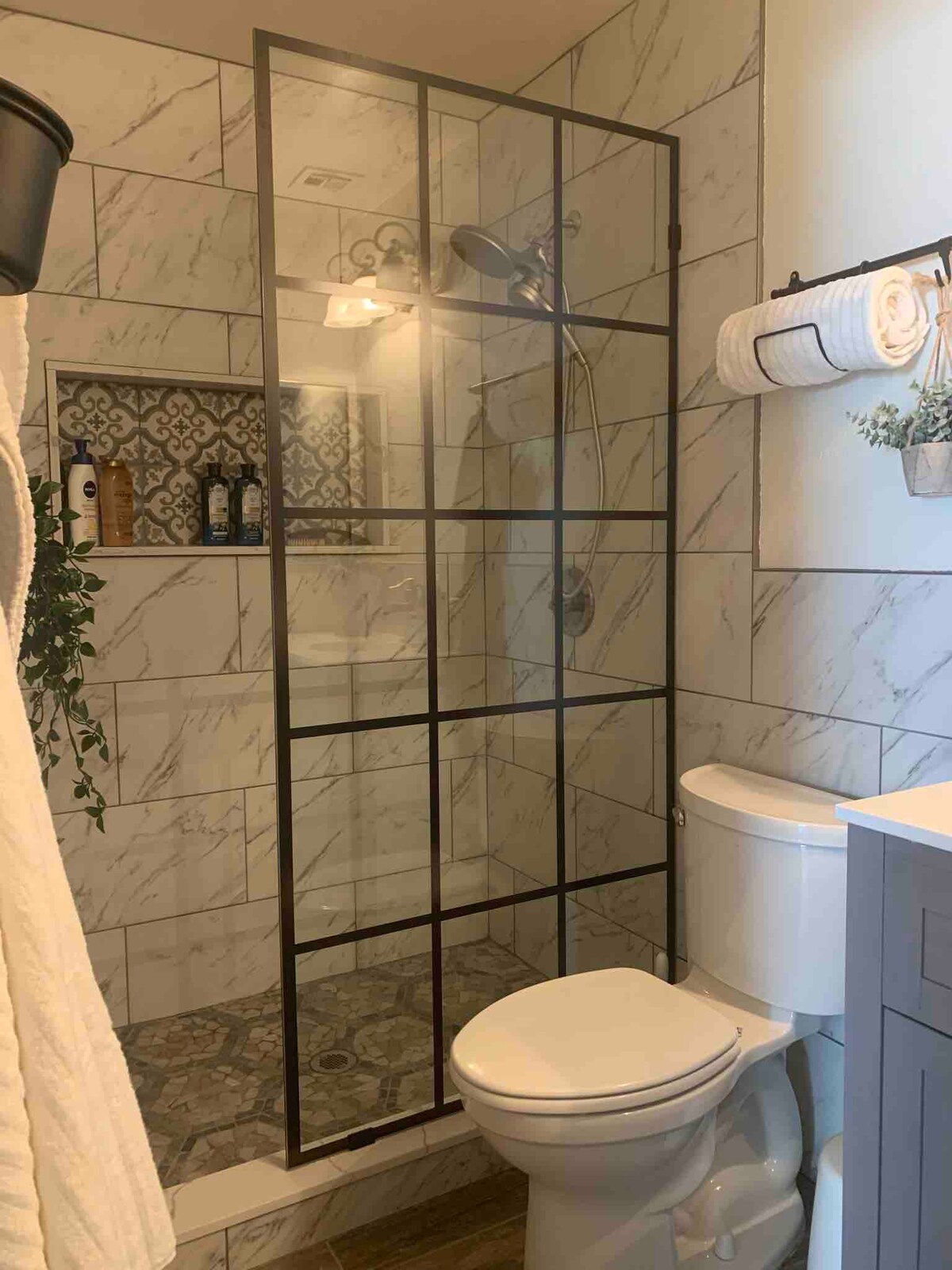 A modern bathroom is presented with a glass shower enclosure featuring a patterned tile backdrop. Natural light shines onto the marble-effect walls, creating a bright environment. A neatly arranged towel rack and decorative elements enhance the space, while a white toilet and gray vanity provide functional aspects.