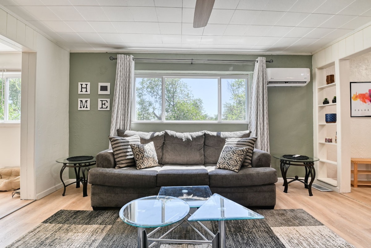A comfortable living area is showcased with a gray couch and decorative cushions. Natural light enters through a large window, while two side tables and a glass coffee table complete the space. Shelving is visible to one side, adding to the room's functionality.