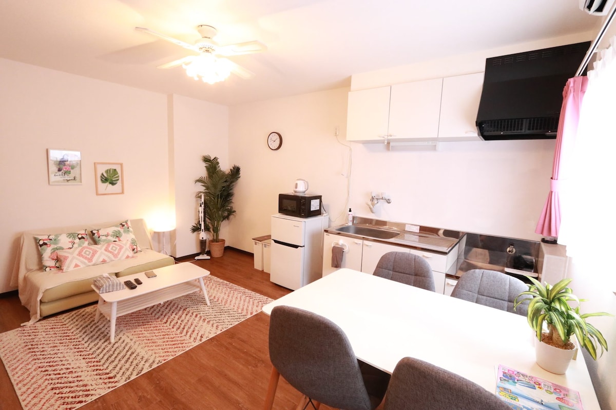 The living area is arranged with a light-colored sofa and a coffee table, complemented by a patterned area rug. A dining table with chairs is positioned nearby. The kitchenette features essential appliances, including a microwave and oven, against a backdrop of simple cabinetry.