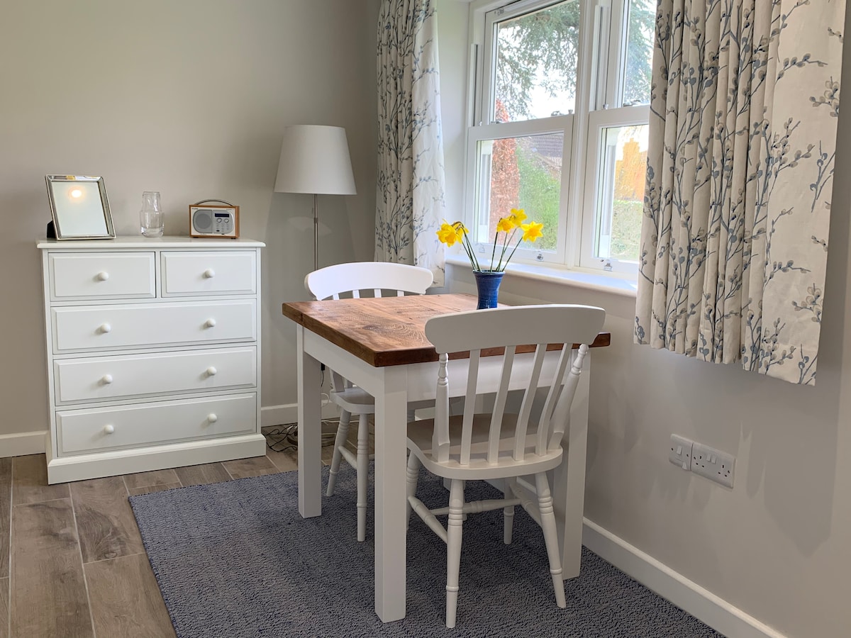 A light-filled sitting area features a wooden table surrounded by two white chairs. A vase of yellow flowers sits on the table, enhancing the scene. Nearby, a chest of drawers and a lamp provide functional elements, and large windows framed with patterned curtains allow natural light to enter.