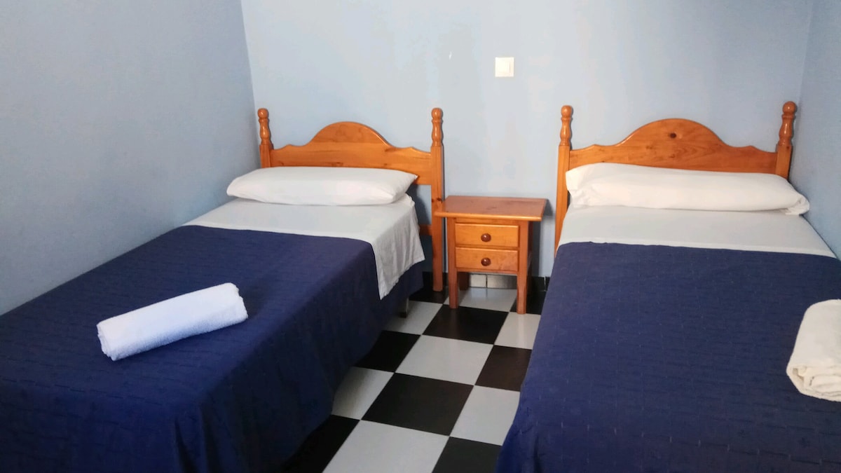 A guest room is presented with two single beds, each topped with white linens and navy blue bedspreads. A small wooden nightstand is positioned between the beds, providing additional space. The floor features a classic black and white checkered pattern, contributing to the room's design.