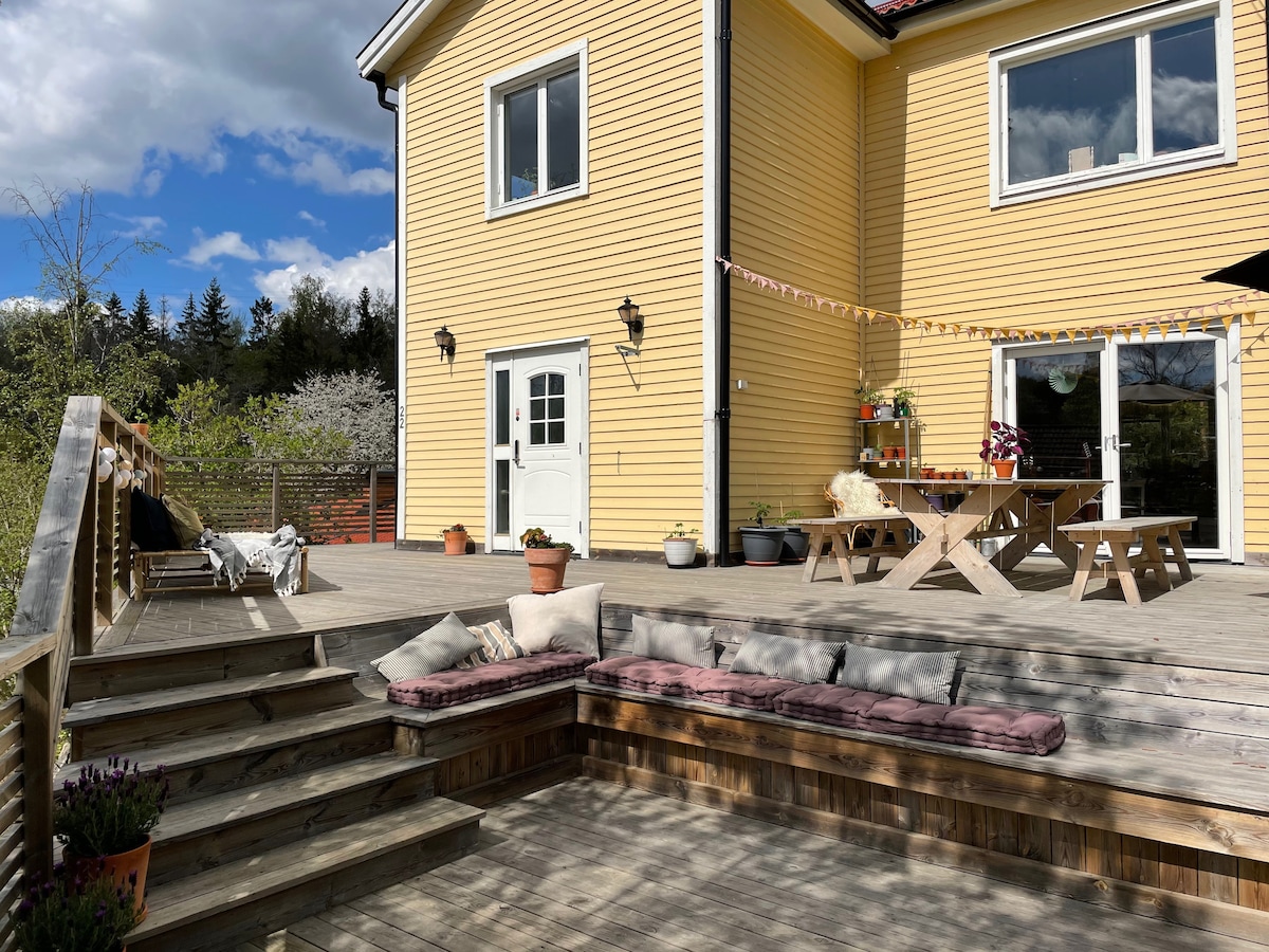 A spacious sun deck is featured in front of a yellow house, complemented by wooden steps leading up to the entrance. Outdoor seating arrangements include a large table and benches, with cushions placed along the deck. Lush greenery and flowering plants create a serene backdrop.