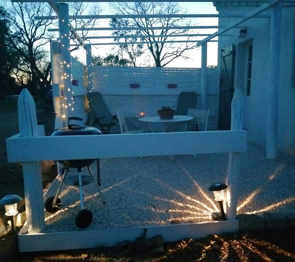 An outdoor patio area is illuminated by soft lighting, highlighting a small table and two chairs. A barbecue grill is positioned nearby, and decorative stones surround the perimeter. A white fence frames the space, complemented by a natural backdrop of trees in the evening light.