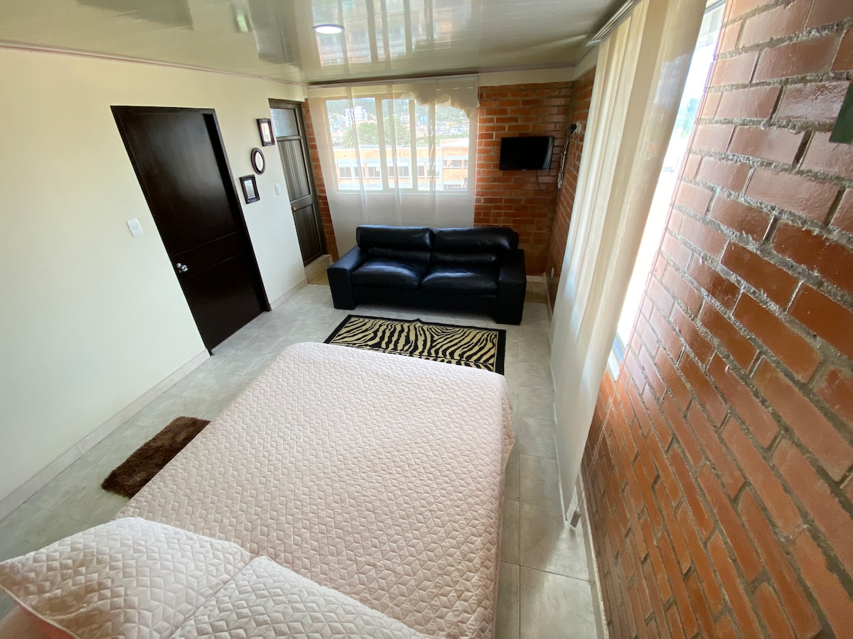 A bright sleeping area is presented with a spacious bed covered in light bedding, while natural light enters through large windows adorned with sheer curtains. A black sofa is positioned in the corner, and a textured area rug complements the design against the neutral tiled floor.
