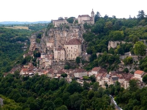A scenic view of a historic village perched on a rocky hillside. Stone buildings with red roofs are nestled among lush greenery, leading up to a prominent hilltop structure. The landscape is dotted with trees, creating a serene backdrop of natural beauty.