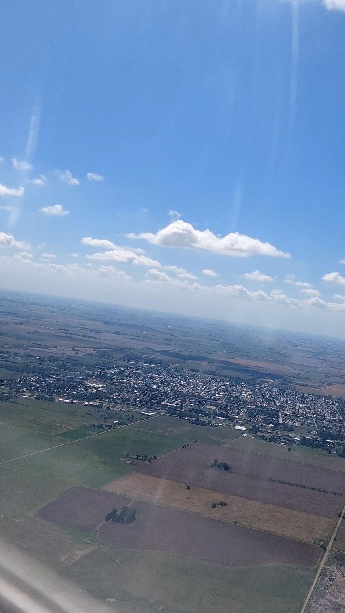 Aerial view captures a sprawling landscape of fields and a small town. The horizon is dotted with fluffy clouds against a bright blue sky, showcasing patches of green and brown land. The layout of the town is visible, with roads and several buildings interspersed among the fields.