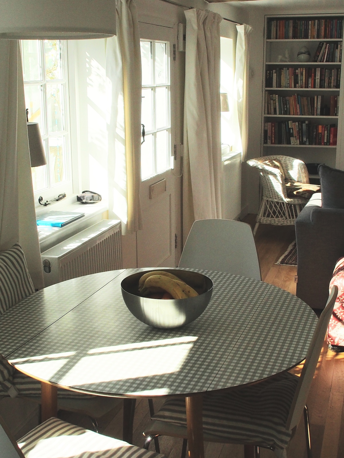 A well-lit dining area features a round table with a bowl of bananas at its center. Surrounding the table are four chairs, and a cozy chair is visible in the background next to a bookshelf filled with books. Light streams through the nearby windows.